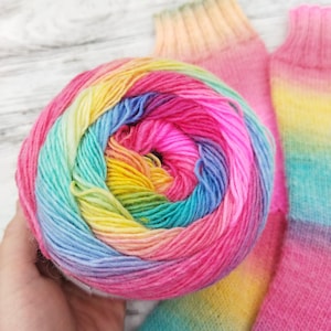 Peut inclure: Une pelote de laine aux couleurs de l'arc-en-ciel, avec des nuances de rose, jaune, bleu et vert. Deux chaussettes partiellement visibles, aux couleurs assorties, sont en arrière-plan. La laine est pour le tricot ou le crochet.