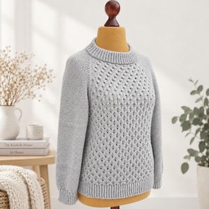 Peut inclure: Un pull à manches longues gris clair présentant un motif en tricot nid d'abeille texturé sur le panneau avant, un tricot uni sur les manches, ainsi qu'un col rond, des poignets et un ourlet côtelés, présenté sur un buste de mannequin.