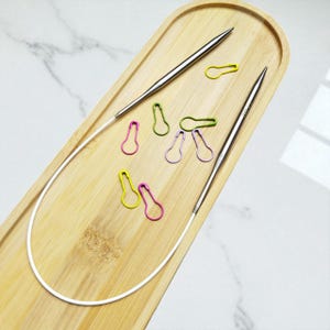 May include: A wooden tray holds a set of silver knitting needles and colorful stitch markers. The needles are curved and connected by a white cable. The stitch markers are in shades of yellow, green, and purple.