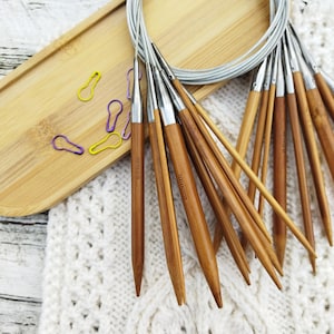 May include: A collection of bamboo circular knitting needles with silver tips, various sizes, and a flexible cable. The needles rest on a wooden tray with colorful stitch markers and a white knitted fabric.
