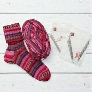 May include: A handmade sock in shades of red, pink, and purple, next to a ball of matching yarn and two packages of knitting needles. The sock features a striped pattern and a detailed cuff. The yarn is a mix of colors.