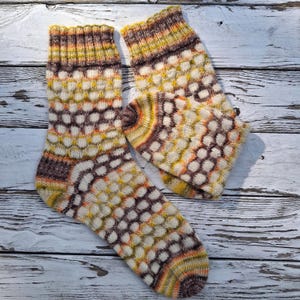 Può includere: Un paio di calze lavorate a maglia a mano con un motivo a nido d'ape nei toni del crema, marrone, giallo e arancione. Le calze presentano un bordo a coste sulla parte superiore e un tallone e una punta rinforzati in filato variegato.
