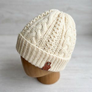 May include: A cream-colored knit beanie featuring a textured cable knit pattern and a ribbed cuff. The hat includes a small, brown, heart-shaped leather tag attached to the front of the cuff with a single silver-toned metal rivet.