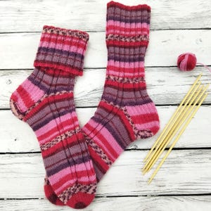 May include: A pair of hand-knitted socks in shades of pink, red, and purple. The socks feature ribbed detailing and a folded cuff. Knitting needles and a ball of yarn are visible, suggesting a handmade craft project.