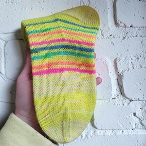 Peut inclure: Une chaussette tricot&eacute;e &agrave; la main avec un motif ray&eacute; color&eacute; et vibrant. La chaussette pr&eacute;sente des bandes horizontales jaunes, roses, oranges, bleues et vertes. Le talon et l'orteil sont de couleur jaune uni. La chaussette est faite d'un fil doux et textur&eacute;.