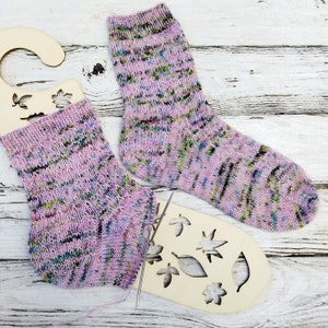 以下が含まれることがあります： ピンクとグリーンの斑点のある靴下2足が木製の表面に置かれています。1足は葉のデザインが施された木製の靴下ブロッカーの上に置かれています。もう1足はブロッカーの横に置かれています。編み針が表面に置かれています。