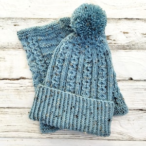 Może przedstawiać: Niebieski zestaw czapki i szalika. Czapka ma prążkowany brzeg i wzorzysty wzór, zwieńczony dużym pomponem. Szalik ma podobny design. Zestaw wykonany jest z miękkiej, nakrapianej przędzy.