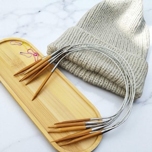 May include: A collection of bamboo circular knitting needles with metal tips, arranged on a light wooden tray. A beige knitted beanie is in the background. Colorful paperclips are also on the tray.