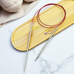 May include: Two silver circular knitting needles with a red cable, displayed on a light brown wooden tray. The needles are pointed and have a smooth, metallic finish. Several colourful paper clips are scattered on the tray, and a ball of yarn is in the background.