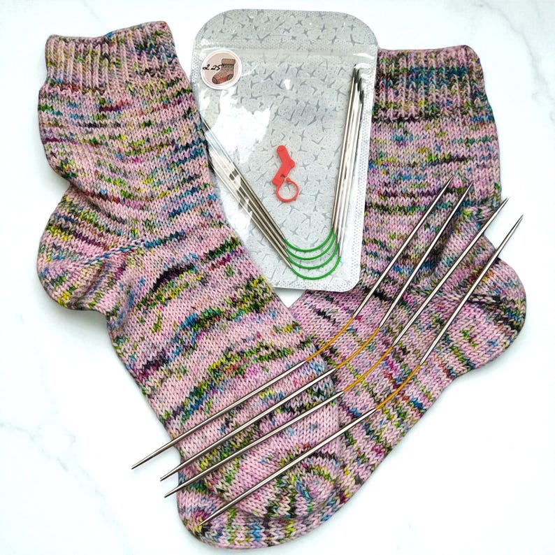 Puede incluir: Un par de calcetines tejidos a mano en hilo jaspeado rosa, verde y azul, con un juego de agujas de tejer y accesorios. La imagen incluye un paquete de pl&aacute;stico transparente con agujas de tejer y marcadores de punto, ideal para tejer.