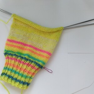 Peut inclure: Une chaussette partiellement tricot&eacute;e en cours, avec un m&eacute;lange de couleurs vives, notamment des rayures jaunes, roses, oranges et bleues. La chaussette est sur des aiguilles &agrave; tricoter en m&eacute;tal, avec du fil jaune sortant du projet. Le projet est sur une surface blanche.