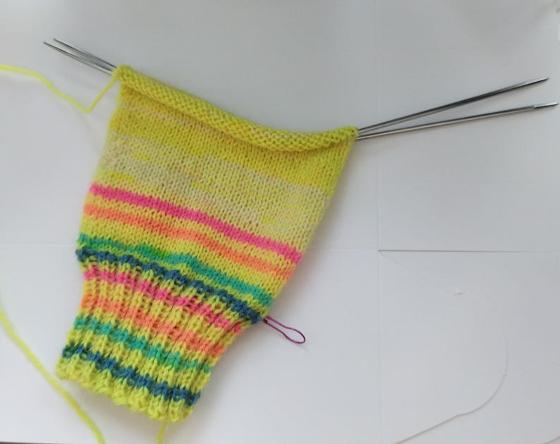 Peut inclure: Une chaussette partiellement tricot&eacute;e en cours, avec un m&eacute;lange de couleurs vives, notamment des rayures jaunes, roses, oranges et bleues. La chaussette est sur des aiguilles &agrave; tricoter en m&eacute;tal, avec du fil jaune sortant du projet. Le projet est sur une surface blanche.