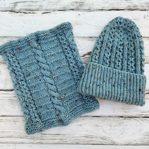 May include: A light blue knitted hat and cowl scarf set. The hat has a folded brim and a textured pattern. The cowl scarf features a cable knit design. Both items have flecks of darker yarn throughout, adding texture and detail.