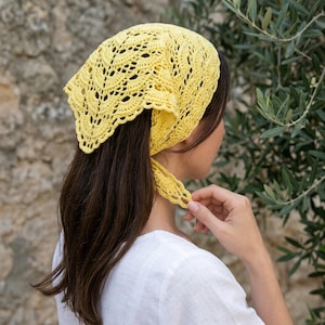 May include: A yellow crochet headscarf with a delicate, openwork design. The head covering is tied at the nape of the neck. The scarf is made of a soft, yellow yarn and is worn over dark brown hair.