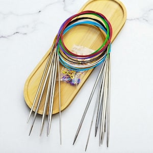 May include: A set of knitting needles with colorful flexible cords, including red, blue, green, and black. The needles are silver and are on a wooden tray, along with a small bag of stitch markers.