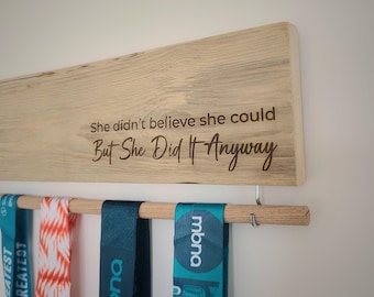 May include: A wooden medal display rack with the inscription "She didn't believe she could But She Did It Anyway". The rack has a wooden dowel with hooks for hanging medals. There are four medals hanging on the rack, two are teal blue with white lettering and two are orange and white.