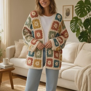 May include: A long-sleeved open-front cardigan made of crocheted granny squares in shades of mustard yellow, sage green, terracotta, and cream. The garment is worn over a plain white t-shirt and light blue denim jeans, featuring a boxy fit.