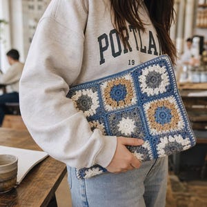 Puede incluir: Una funda rectangular de ganchillo para portátil construida con cuadrados de la abuela unidos en tonos azul, beige, gris y blanco. La funda presenta un patrón floral circular repetitivo en cada cuadrado, enmarcado por bordes azules, y se sostiene contra una sudadera gris claro.