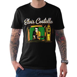 May include: Black t-shirt with a vintage television graphic featuring Elvis Costello. The television screen shows Costello playing a guitar in front of a green and black checkered background. The text "Elvis Costello" is written in white with a yellow outline.