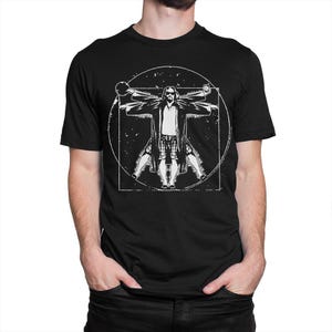 May include: A black t-shirt featuring a white graphic of a man in a robe and shorts, posed like the Vitruvian Man, with the text "The Dude Abides".