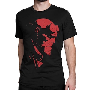 Puede incluir: Una camiseta negra con una silueta roja de la cabeza del personaje Hellboy. La cara del personaje es visible, con una cabeza roja con cuernos y un solo ojo.