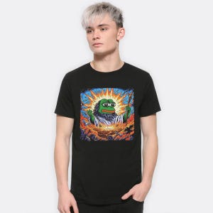 Puede incluir: Camiseta negra con un estampado gráfico de una rana verde con halo y un bastón, de pie frente a un fondo naranja y amarillo brillante con un sol y nubes.