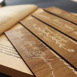 Puede incluir: Primer plano de marcadores de libros de madera con nombres y fechas grabados, con dise&ntilde;os florales. Los marcadores son de color marr&oacute;n c&aacute;lido y est&aacute;n colocados sobre un libro abierto. El texto incluye nombres y fechas.