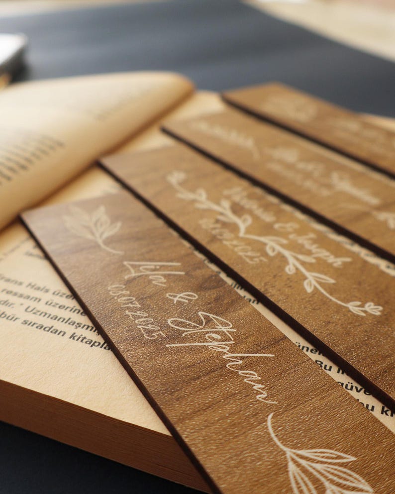 Puede incluir: Primer plano de marcadores de libros de madera con nombres y fechas grabados, con dise&ntilde;os florales. Los marcadores son de color marr&oacute;n c&aacute;lido y est&aacute;n colocados sobre un libro abierto. El texto incluye nombres y fechas.