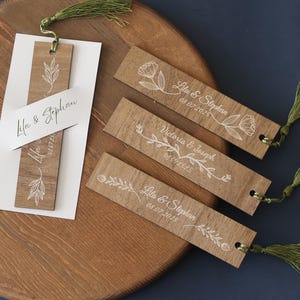 Puede incluir: Marcadores de libros de madera con dise&ntilde;os florales blancos y nombres con fechas. Los marcadores tienen borlas verdes y est&aacute;n sobre una bandeja de madera. El texto en los marcadores incluye nombres como "Lila & Stephan" y fechas como "08.07.2025".