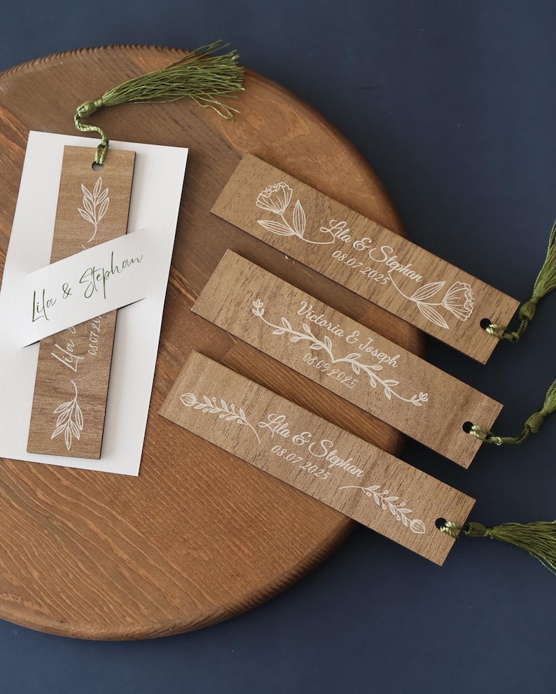 Puede incluir: Marcadores de libros de madera con dise&ntilde;os florales blancos y nombres con fechas. Los marcadores tienen borlas verdes y est&aacute;n sobre una bandeja de madera. El texto en los marcadores incluye nombres como "Lila & Stephan" y fechas como "08.07.2025".