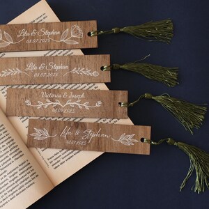 Puede incluir: Marcadores de madera con dise&ntilde;os florales blancos y texto personalizado, incluyendo nombres y fechas. Cada marcador tiene una borla verde. Los marcadores se muestran en un libro abierto sobre un fondo azul oscuro.
