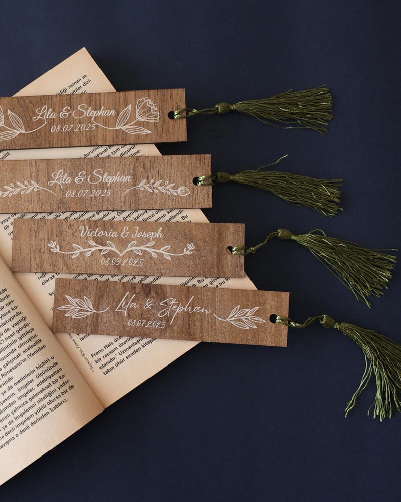 Puede incluir: Marcadores de madera con dise&ntilde;os florales blancos y texto personalizado, incluyendo nombres y fechas. Cada marcador tiene una borla verde. Los marcadores se muestran en un libro abierto sobre un fondo azul oscuro.