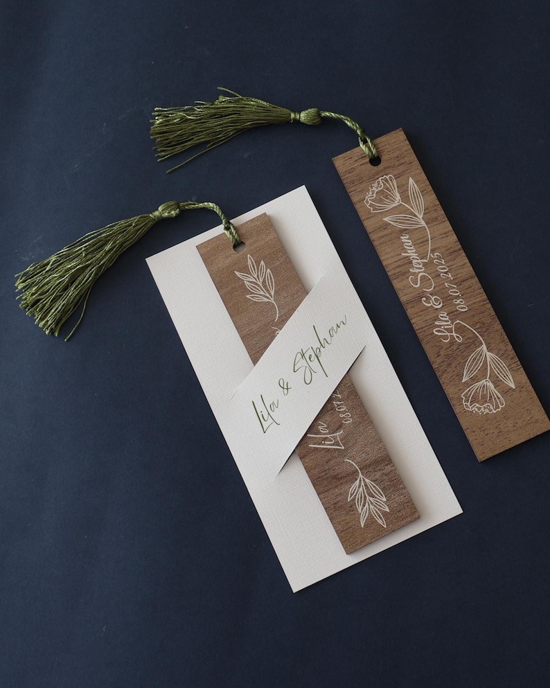 Puede incluir: Marcadores de libros de madera con dise&ntilde;os florales blancos y borlas verdes. Un marcador presenta los nombres "Lila & Stephan" y la fecha "08.07.2025". Los marcadores est&aacute;n sobre una tarjeta blanca, sobre fondo azul oscuro.
