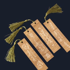 Puede incluir: Cuatro marcadores de madera con dise&ntilde;os florales blancos y nombres con fechas. Cada marcador tiene una borla dorada adjunta. Los marcadores est&aacute;n dispuestos en diagonal sobre un fondo azul oscuro.