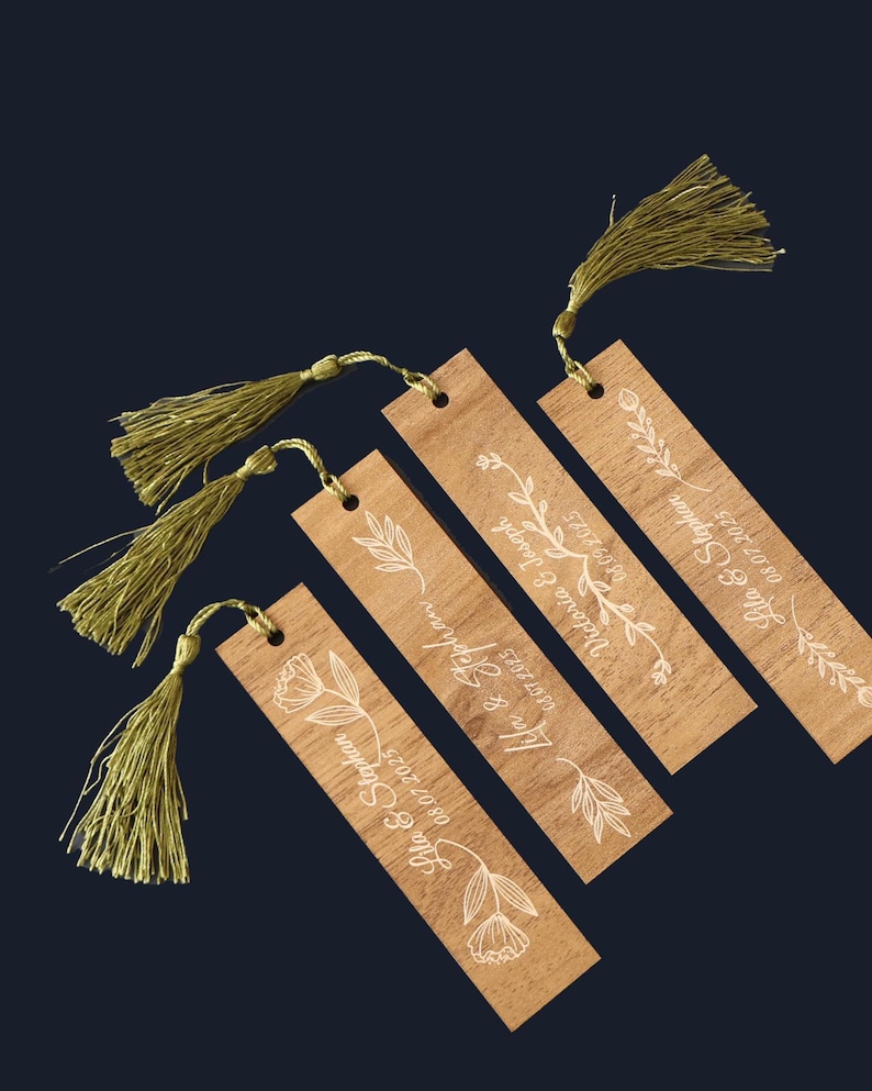 Puede incluir: Cuatro marcadores de madera con dise&ntilde;os florales blancos y nombres con fechas. Cada marcador tiene una borla dorada adjunta. Los marcadores est&aacute;n dispuestos en diagonal sobre un fondo azul oscuro.
