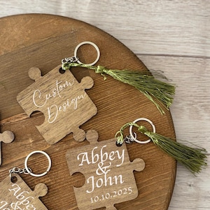 Puede incluir: Llaveros de madera con forma de pieza de rompecabezas con llaveros plateados y borlas verdes. Los llaveros están grabados con nombres y fechas en escritura blanca. Un llavero dice "Custom Design". Otro dice "Abbey & John 10.10.2025".