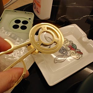 May include: A gold-colored, decorative scissor-like tool with a circular design. The tool is held near a white square dish with a colorful illustration. A light green phone case is in the background.