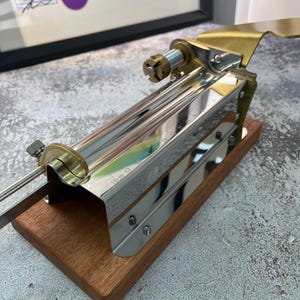 May include: A knife sharpening tool with a wooden base, stainless steel body, and brass accents. The tool has a sliding mechanism with measurement markings and a blade guide. The base is made of dark wood. The tool is designed for precision sharpening.