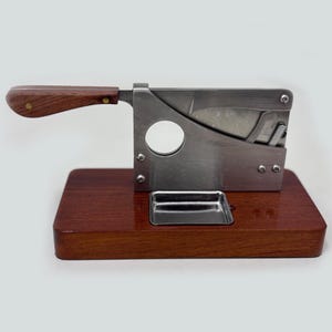 May include: A cigar cutter with a stainless steel blade and a wooden handle. The cutter is mounted on a rectangular, polished, dark wood base. A small, stainless steel tray is attached to the base.