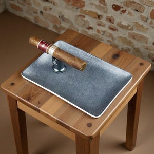 以下が含まれることがあります： 茶色の葉巻が、白い縁取りのある濃い灰色の長方形のトレイの上に置かれ、銀色の葉巻スタンドで支えられています。 トレイは、レンガの壁を背景にした小さな木製のテーブルの上にあります。