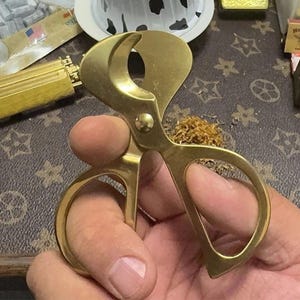 May include: A gold-colored cigar cutter with a circular blade and scissor-like handles. The cutter is held in a hand, with a small pile of tobacco visible nearby. The background features a patterned surface and other accessories.