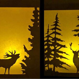 Könnte beinhalten: Zwei beleuchtete Papierauschnitte, die Waldszenen mit Hirschen darstellen. Eines zeigt einen Hirsch mit großem Geweih, silhouettiert gegen einen warmen gelben Schein, umgeben von Bäumen. Das andere zeigt einen Hirsch, der in einem Wald mit hohen Bäumen steht.