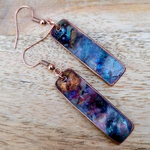 Może przedstawiać: Para prostokątnych kolczyków z miedzianym wykończeniem i kolorowym, opalizującym wzorem. Kolczyki mają zapięcie na haczyk i niepowtarzalny, artystyczny wygląd. Kolory obejmują odcienie niebieskiego, fioletu i złota.