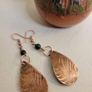 May include: Copper teardrop earrings with etched patterns and dark green beads. The earrings have a rustic, handcrafted look. A small, brown ceramic pot with green and black details is in the background.