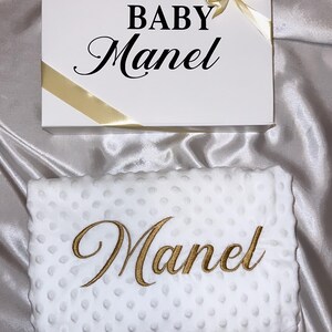 Peut inclure: Une boîte cadeau blanche avec un ruban doré et le texte "BABY Manel" sur le dessus. Une couverture blanche avec une broderie dorée qui dit "Manel" est à côté de la boîte.