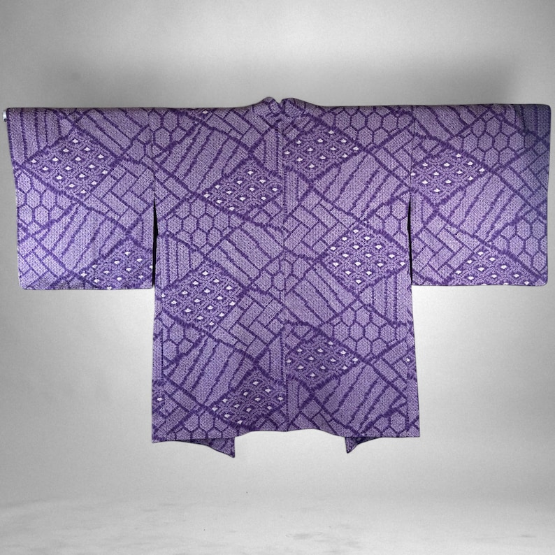 Puede incluir: Un kimono morado con un patr&oacute;n geom&eacute;trico. El kimono presenta un dise&ntilde;o repetitivo de rect&aacute;ngulos, hex&aacute;gonos y rombos. La tela tiene una apariencia texturizada, y la est&eacute;tica general es tradicional y elegante.
