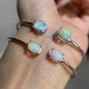 Puede incluir: Cuatro pulseras de puño abiertas con piedras preciosas de ópalo ovaladas. Dos pulseras son de metal plateado y dos de oro rosa. Los ópalos muestran un juego de colores con destellos verdes, azules y amarillos.