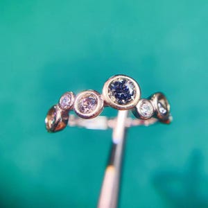 May include: A silver ring with a row of five round clear and grey gemstones. The ring is on a blue background.