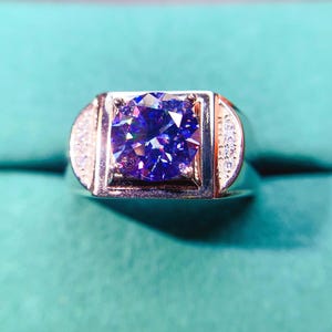 Könnte beinhalten: Ein silberner Ring mit einem großen, runden, violetten Edelstein, der von kleineren, weißen Edelsteinen umgeben ist.