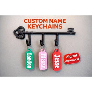 May include: A black metal key rack with three custom name keychains. The keychains are green, pink, and red, each with a different name in white. The text "CUSTOM NAME KEYCHAINS" is above the key rack.